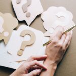 Photo of paper question marks with hands and a pen suggesting curiosity and inquiry.