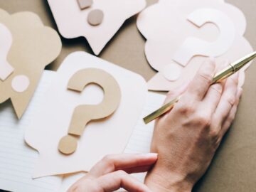 Photo of paper question marks with hands and a pen suggesting curiosity and inquiry.