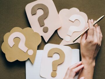 Hands holding pen next to paper question marks on notebook, symbolizing inquiry and creativity.