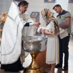 A priest performs a baptism ceremony on a baby in a church, witnessed by parents.