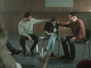Three men in a gymnasium engaged in a supportive group therapy session.