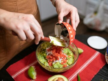 A person prepares homemade guacamole with avocado, tomato, and lime in a kitchen setting.