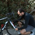 A man kneels to repair a bicycle tire, surrounded by lush greenery, illustrating outdoor activity.