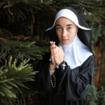 A nun in traditional habit holds a rosary near a tree, symbolizing faith and devotion.