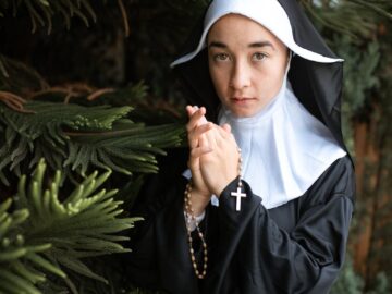 A nun in traditional habit holds a rosary near a tree, symbolizing faith and devotion.