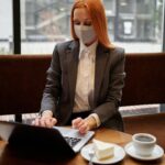Professional woman in a mask works on a laptop in a cafe, embodying modern remote work trends.