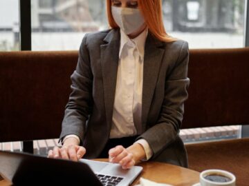 Professional woman in a mask works on a laptop in a cafe, embodying modern remote work trends.