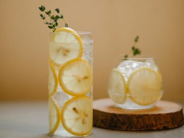 A cool, refreshing drink with lemon slices and thyme in clear glasses on a wooden coaster.