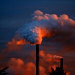 Smoke billows from industrial chimneys at sunset, highlighting pollution against a vibrant sky.