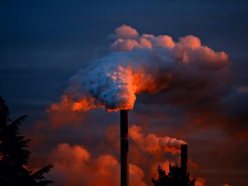 Smoke billows from industrial chimneys at sunset, highlighting pollution against a vibrant sky.