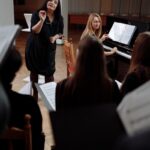 A choir rehearsal indoors with a conductor leading and a pianist accompanying.
