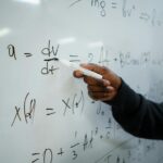 Close-up of hand writing equations on a whiteboard in a classroom setting.