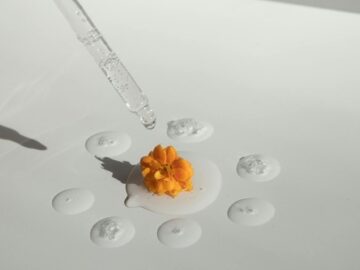 From above of drops of transparent moisturizing cosmetic product dripped by pipette and small fresh flower head placed on white table