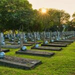 Rows of tombstones with military helmets located on grassy ground near tall lush green trees in heroes cemetery in Kalibata