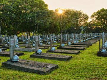 Rows of tombstones with military helmets located on grassy ground near tall lush green trees in heroes cemetery in Kalibata
