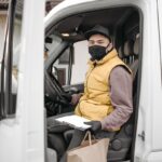 A delivery driver wearing a face mask sits inside a van holding a paper bag and clipboard, symbolizing contactless delivery