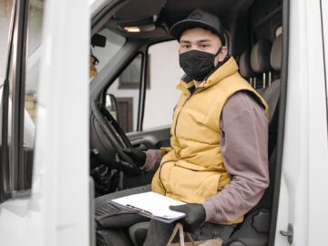A delivery driver wearing a face mask sits inside a van holding a paper bag and clipboard, symbolizing contactless delivery