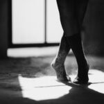 Black and white of crop anonymous female ballet dancer standing on dusty floor with crossed legs