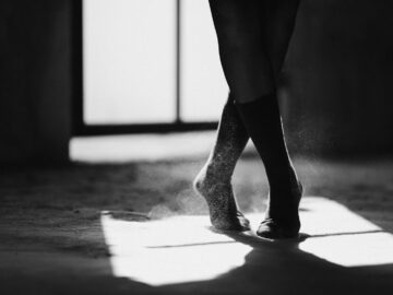 Black and white of crop anonymous female ballet dancer standing on dusty floor with crossed legs