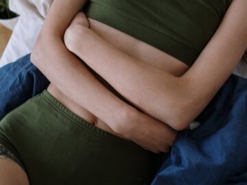 A woman in green clothing lies on a bed clutching her abdomen, suggesting pain or discomfort.