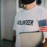 Volunteer wearing a mask holds a notebook and American flag, symbolizing community service.