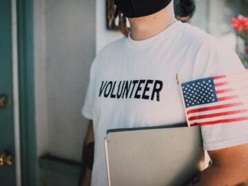 Volunteer wearing a mask holds a notebook and American flag, symbolizing community service.