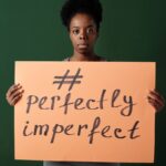 African American woman holding a sign that reads '#perfectly imperfect' on a green background.