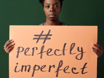 African American woman holding a sign that reads '#perfectly imperfect' on a green background.