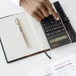 A hand using a calculator with financial documents and notebook, depicting an office setup for financial analysis.