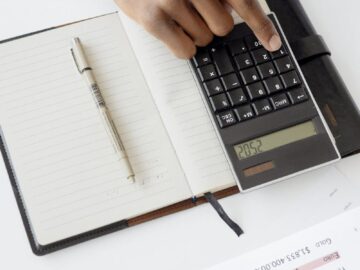 A hand using a calculator with financial documents and notebook, depicting an office setup for financial analysis.