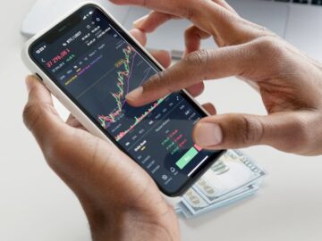 Hands using smartphone trading app with charts, dollar bills in background.