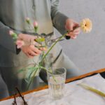 A woman in green outfit trimming flowers for a floral arrangement indoors.