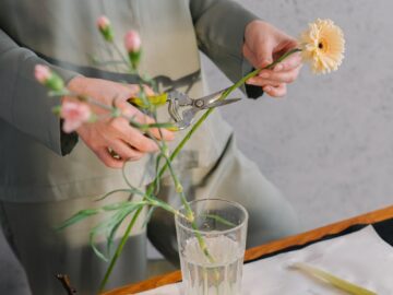 A woman in green outfit trimming flowers for a floral arrangement indoors.
