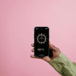 Close-up of a hand holding a smartphone displaying a compass app against a pink background.