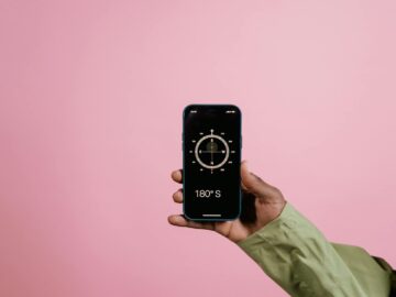 Close-up of a hand holding a smartphone displaying a compass app against a pink background.