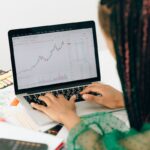 A woman working on financial analysis using a laptop with a stock market graph on screen.