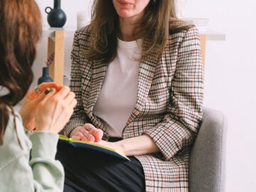 Serious adult female therapist in elegant outfit sitting in armchair and listening to crop anonymous patient talking about problems during psychotherapy consultation in light office