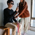A woman supports a visually impaired man indoors, highlighting compassion and assistance.