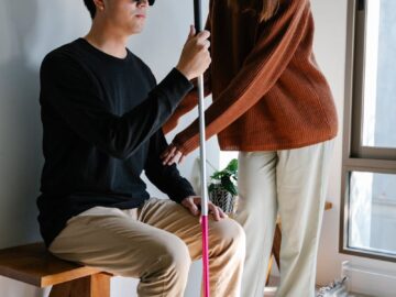 A woman supports a visually impaired man indoors, highlighting compassion and assistance.