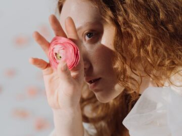 Artistic portrait of a redheaded woman holding a pink flower over one eye.