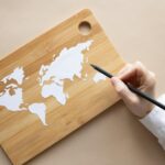Hand crafting a world map on a bamboo cutting board using a pencil on beige background.