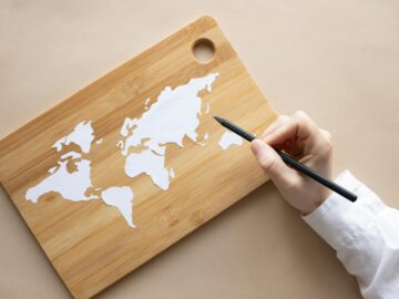 Hand crafting a world map on a bamboo cutting board using a pencil on beige background.