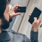 Adult man using a smartphone for online transactions while holding a credit card indoors.