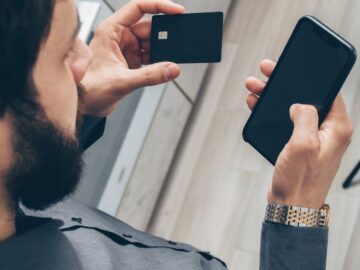 Adult man using a smartphone for online transactions while holding a credit card indoors.