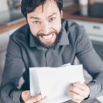 A frustrated man holding papers in a kitchen, showing stress and anxiety.