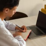 A young man focusing on his studies at home, using a laptop and notebook.