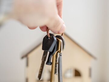 Close-up of a hand holding keys with a miniature wooden house in the background, symbolizing real estate investment.
