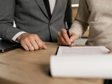 Close-up of businessmen signing documents at a wooden table in an office.