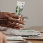 Close-up of hands counting US dollar bills on a cluttered desk in an office setting.