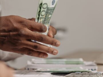 Close-up of hands counting US dollar bills on a cluttered desk in an office setting.